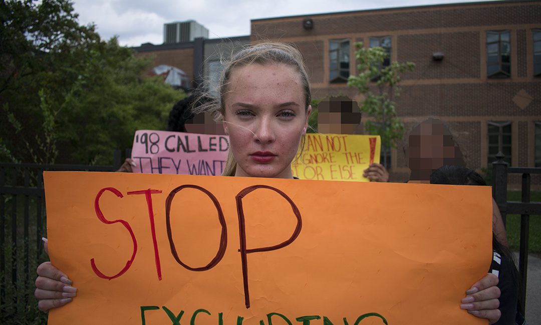 Quinne Kendall-Mitchell, a grade 10 student at East York Collegiate Institute, stands in front of her school, holding a yellow sign which reads "Stop excluding LGBTQ."