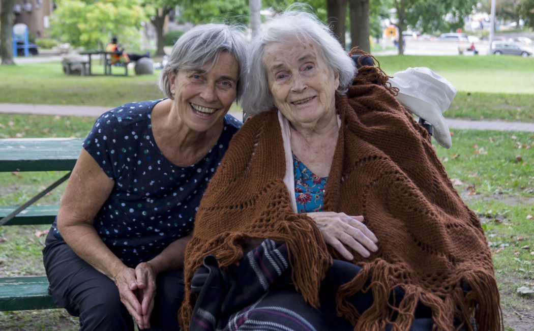 Catherine Staples and Mary Staples (she is in a wheelchair) are smiling for the camera in a park before talking about their lives in East York.
