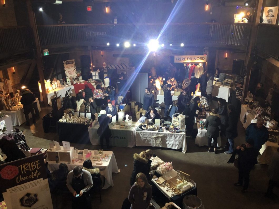 Shoppers look at goods including jewelry, food, and art in a dimly lit space with one shining bright light in the top centre.