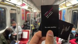 Hands holding up two business cards with the words "I worry that we're not meant for one another" and "Commuting and Other Lonely Thoughts" written on them. Held up in a Toronto subway filled with people sitting by themselves