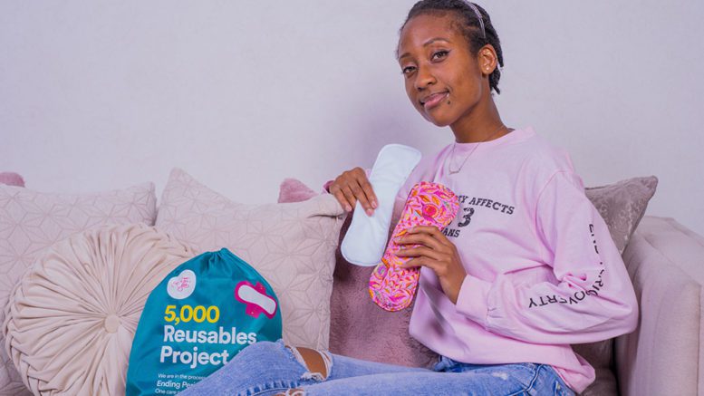 Help a Girl Out volunteer sitting on a couch holding two of the reusable pads sewn for the 5,000 reusables project, with a period product kit propped up with pillows to the left of her.