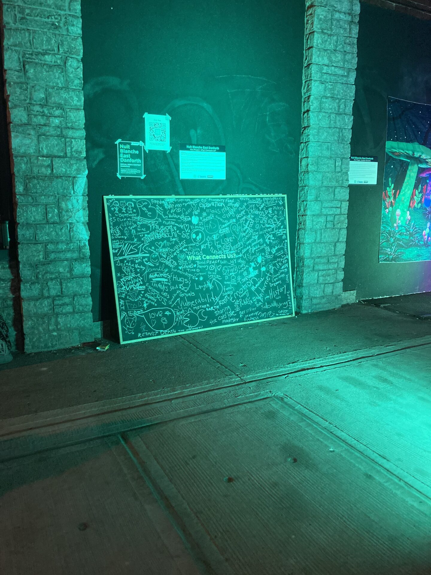 A fluorescent turquoise light shines against a chalkboard, covered in words and drawings related to the theme of "What Connects Us."