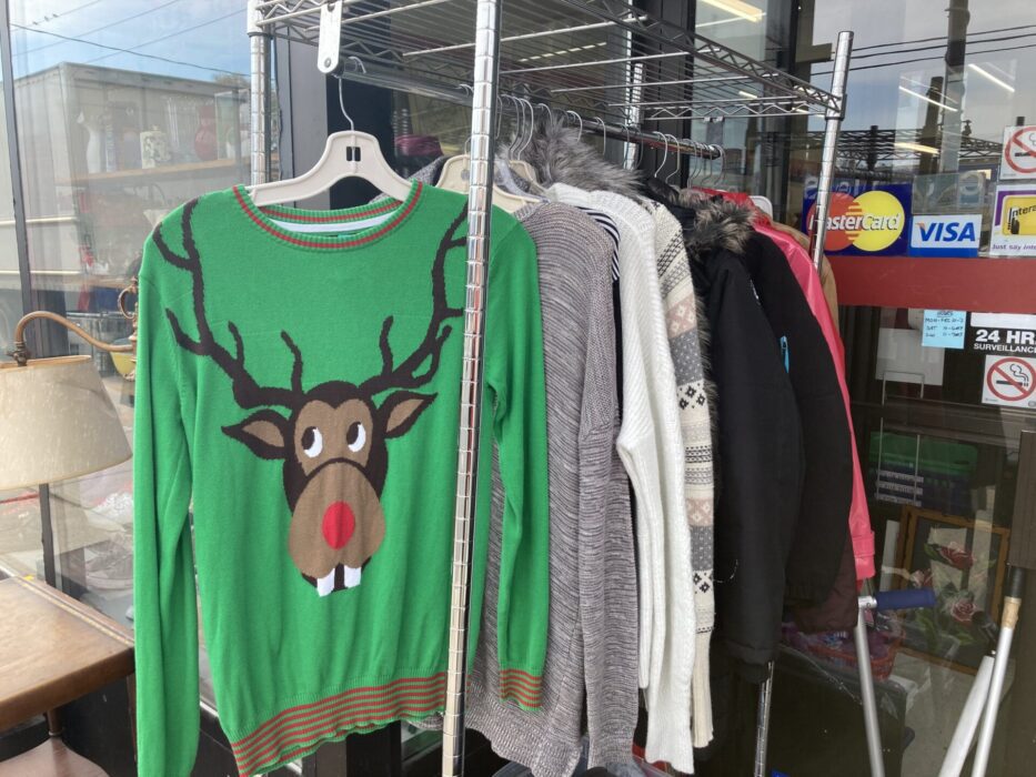 A green sweater with Rudolph the red-nosed reindeer printed on the front, on a rack of various sweaters outside a thrift store