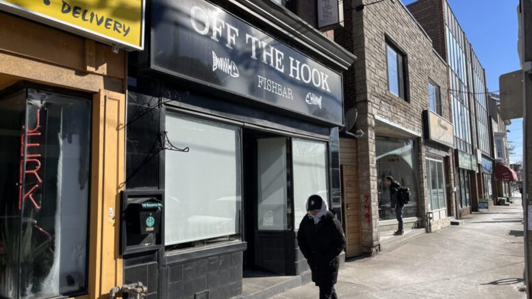 A Toronto citizen walks in front of the restaurant Off the Hook in East York. Doors closed and curtains down, with a big black sign receiving light from a sunny and yet cold day.