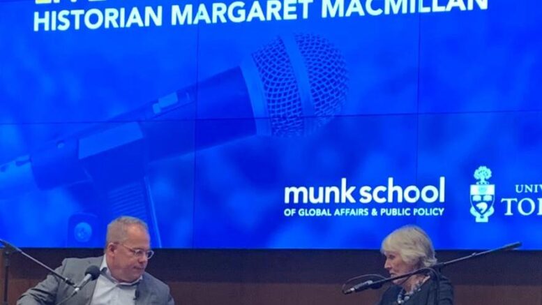 Paul Wells and Margaret MacMillan sit across each other, talking, in front of a blue screen reading "Paul Wells Show Live Podcast Taping: Historian Margaret MacMillan."