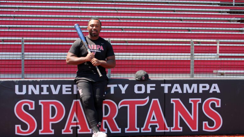 Jamarcus Lyons holds his bat in front of the Tampa University Spartans logo behind home plate.