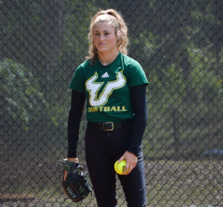 Pitcher Payton Dixon waits for instructions from catcher wearing her team shirt with a ball and glove in her hands
