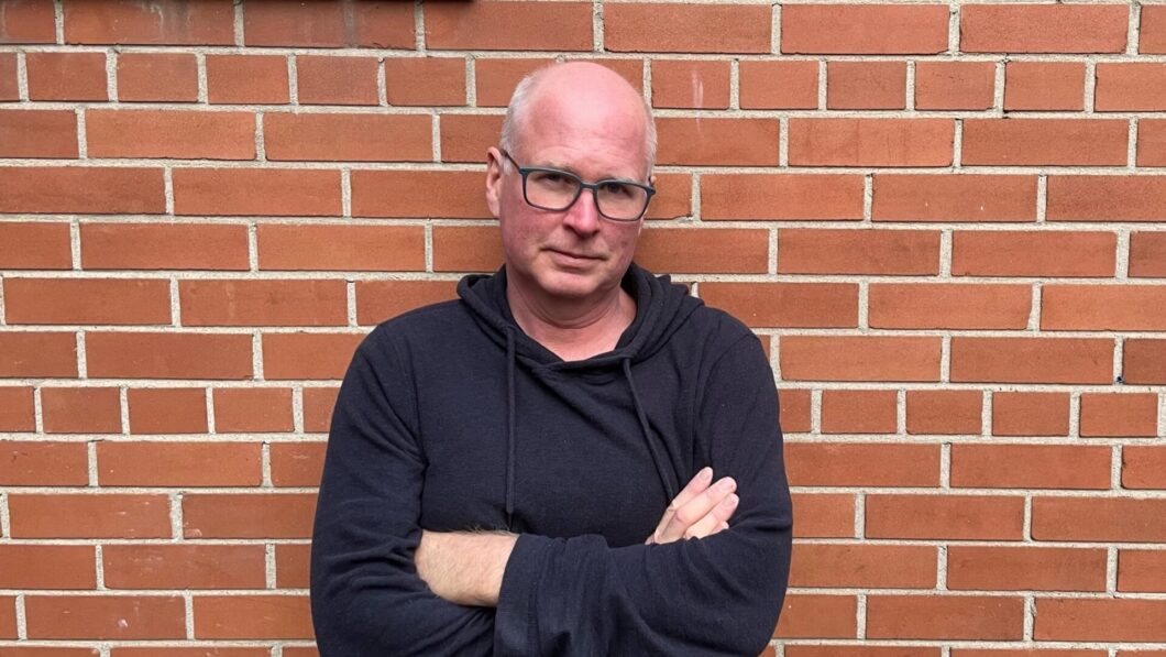 Journalist Dave Seglins stands in front of a brick wall with his arms crossed.