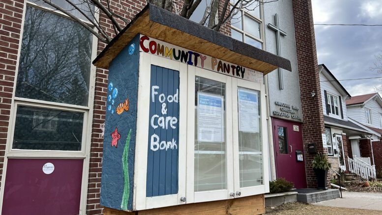A wooden community pantry in front of Kimbourne Park United Church. The words 'community pantry' written at the top of the pantry and 'food & care bank' written on the left pantry door. A fish blowing bubbles and a starfish are drawn on the left side of the pantry.