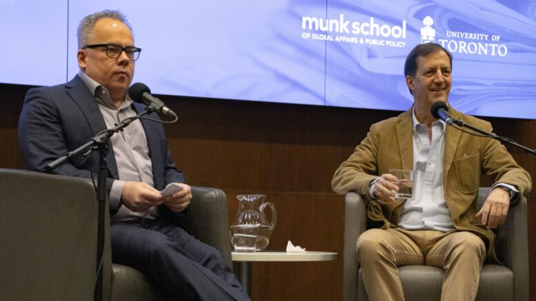 Paul Wells and Andrew Coyne facing the audience. Coyne smiling and holding a glass of water. Microphones in front of them. About to speak for The Paul Wells Show interview podcast.