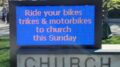 A blue sign outside St. Aidan's church that says "Ride your bikes, trikes & motorcycles to church this Sunday"